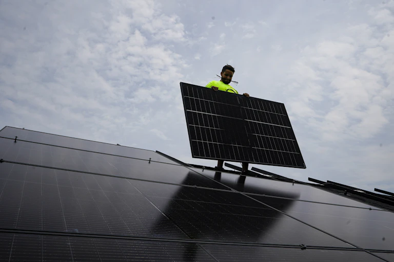 A man holds up a solar panel on a roof against a cloudy sky.