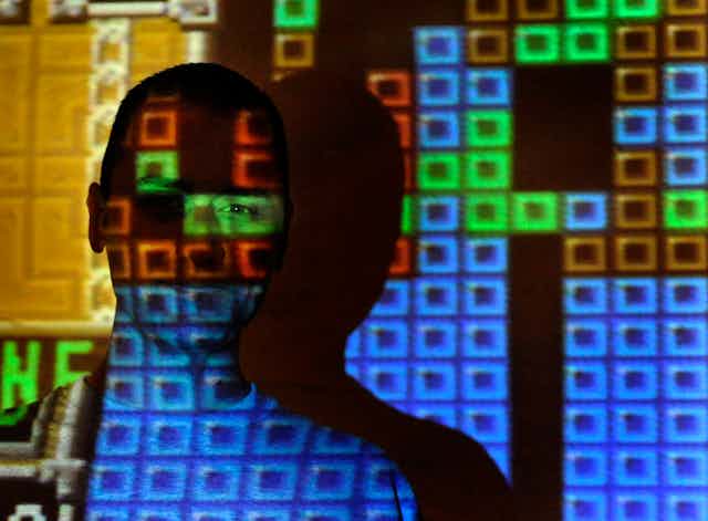 Projection of pixelated, colored blocks beams across a teenaged boy standing in front of a screen.