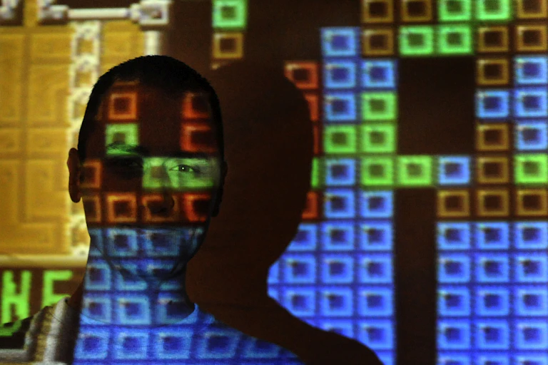 Projection of pixelated, colored blocks beams across a teenaged boy standing in front of a screen.