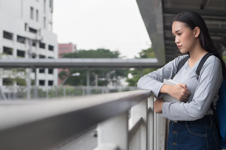 An asian woman holding a book looks off into the distance with a worried look on her face