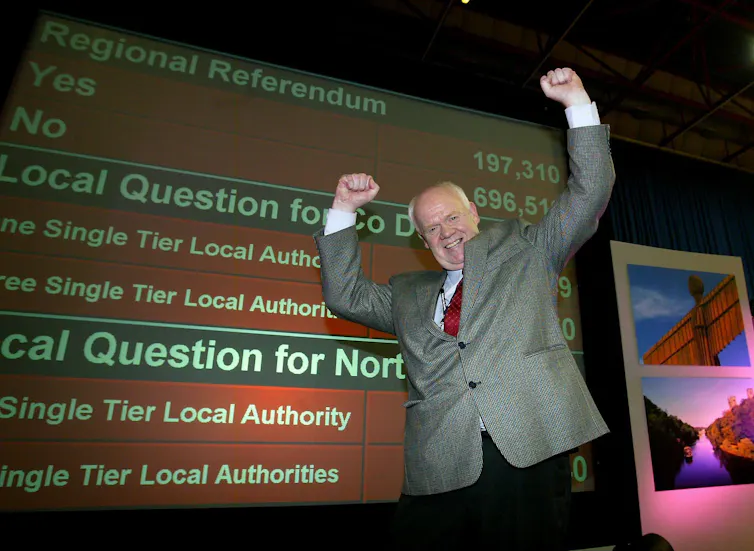 Grey-haired man celebrates with arms aloft in front of an election sign
