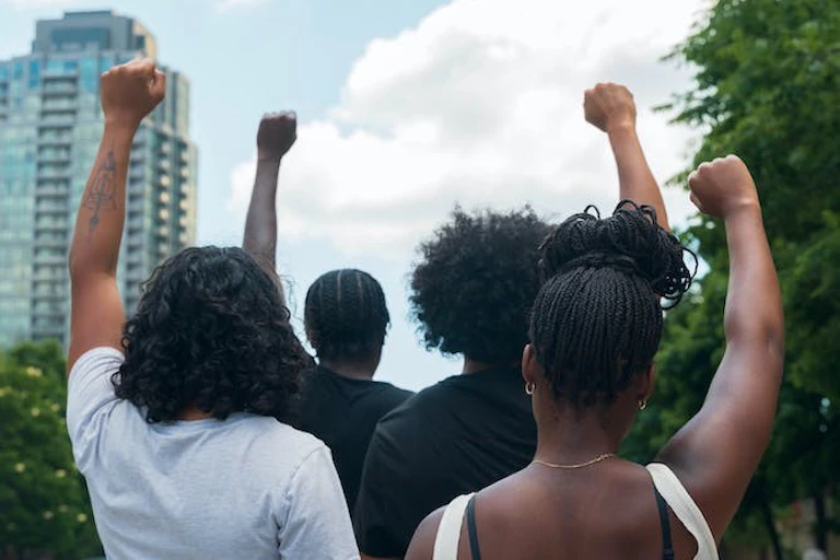 Deux hommes et deux femmes lèvent le poing en signe de protestation, dos à la caméra et face à une tour d'habitation.
