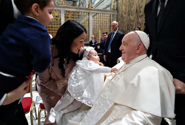 Pope Francis holds a baby dressed in white, while the family looks on.