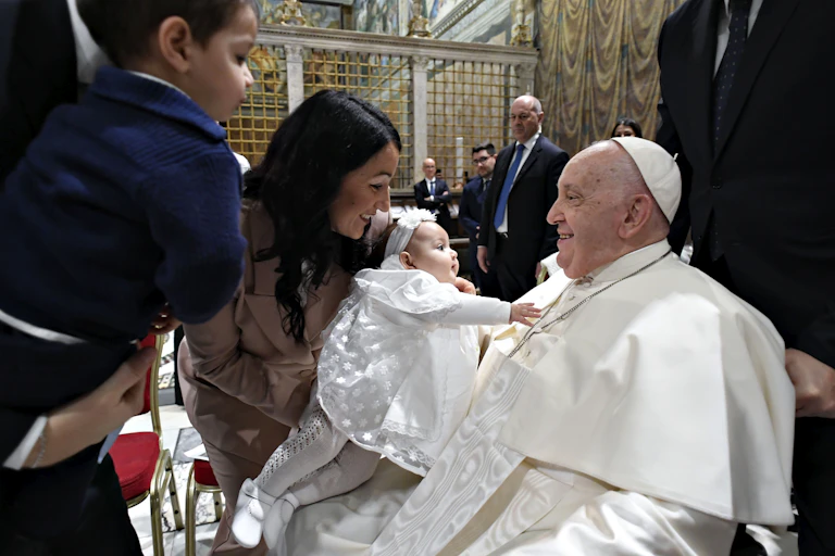 Pope Francis holds a baby dressed in white, while the family looks on.