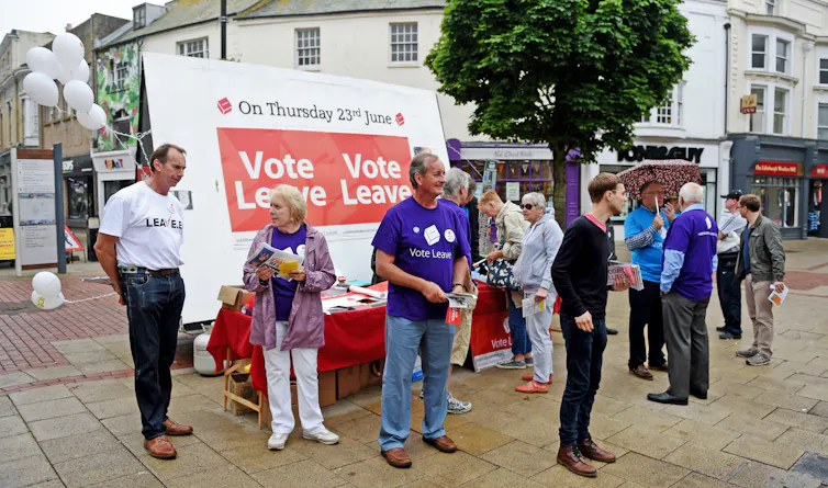 People stand on a street campaigning for Brexit votes with a 'Vote Leave' banner behind them.