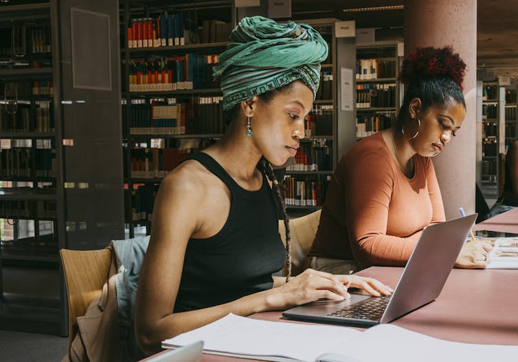 Two young Black women work from a desk at a library.