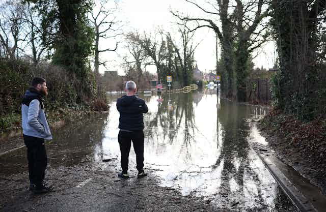 Two men stand at the edge of a submerged road outside a village.