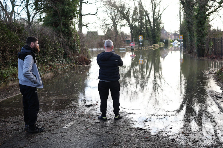 Two men stand at the edge of a submerged road outside a village.