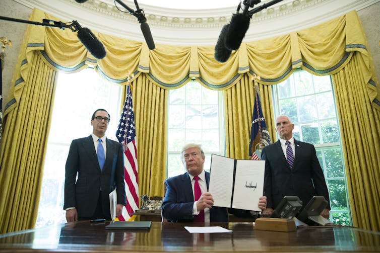 A man in a suit, seated at a desk, holding up a signed document and flanked by two other men in suits who are standing.