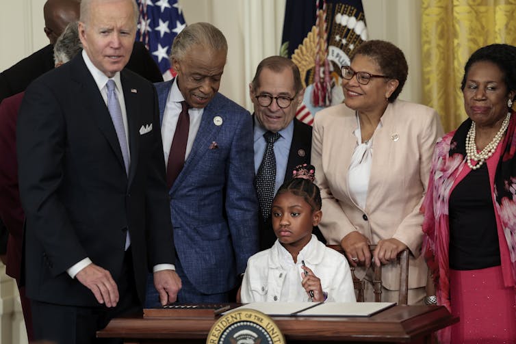 A young girl sits at a table holding a pen, surrounded by adults.