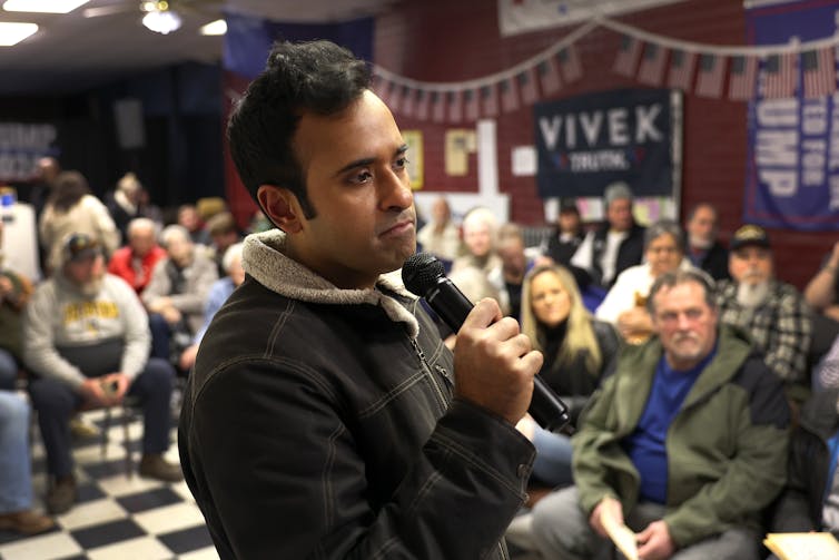 A brown-skinned man holds a microphone as dozens of white people listen to his campaign speech.