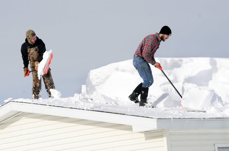 Two men shovel snow off a roof. The snow is is higher than their waists.