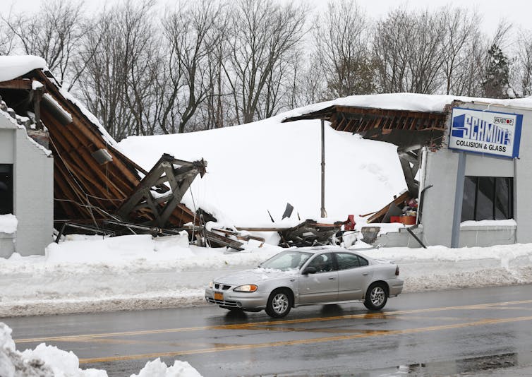 A vehicle drives past a show with the roof bowed all the way to the ground with a thick layer of snow on top.
