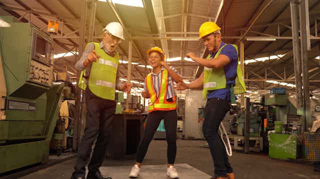 Three people in high-vis vests and hard hats dance, factory in the background.