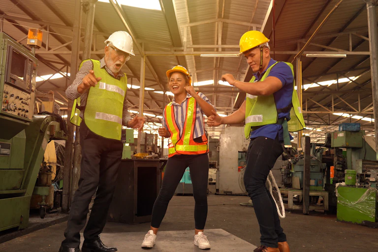 Three people in high-vis vests and hard hats dance, factory in the background.