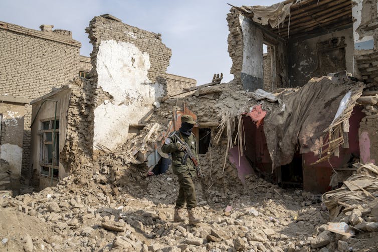 A man in fatigues stands on rubble, broken walls are behind him.