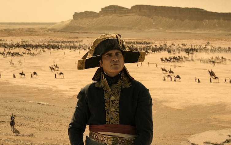 A man wearing a bicorne hat and a single-breasted blue coat with gold detailing in front of a desert landscape