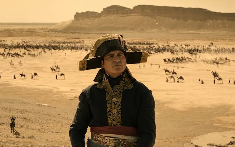 Un homme portant un chapeau bicorne et un manteau bleu à simple boutonnage avec des détails dorés devant un paysage désertique