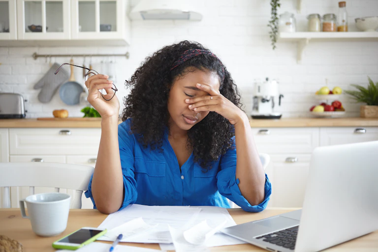 A woman working on a laptop at a kitchen table. She holds her head in her hand.