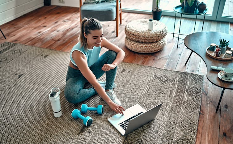 A woman wearing exercise clothes sits on a floor using a laptop. Dumbbells are on he floor beside her.