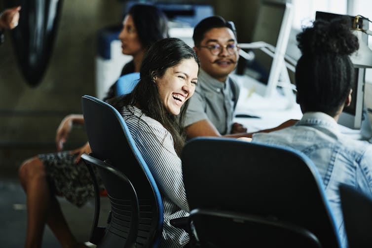 A woman seated on a chair, with two other coworkers, laughs while having a conversation.