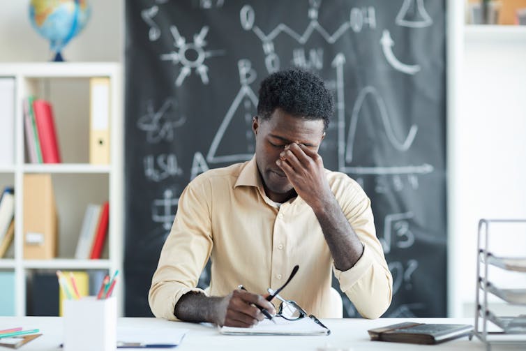 A young Black teacher, looking tired, at his desk in a classroom.