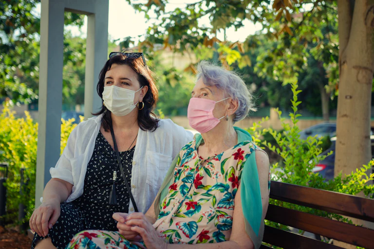 A younger woman and an older woman wearing masks sitting on a bench.