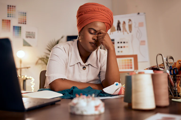 Woman at desk at home, surrounded by dressmaking equipment, eyes closed, hand to forehead.