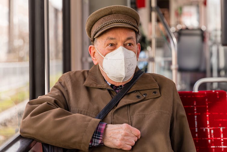 Older male seated on a bus, wearing a brown coat, hat and white COVID mask
