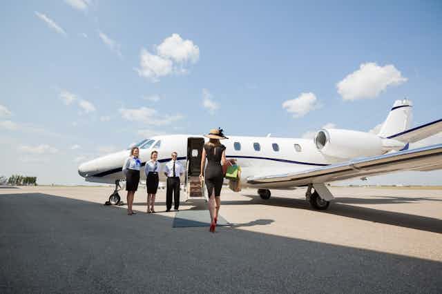 A rich woman walks towards the entrance of a private jet with three crew members standing outside.