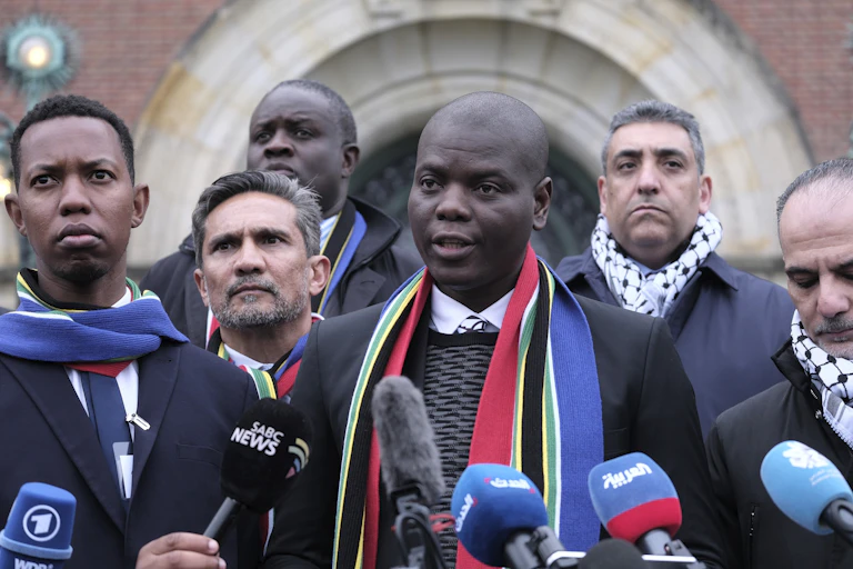A Black man speaks behind a row of microphones as other men surround him.