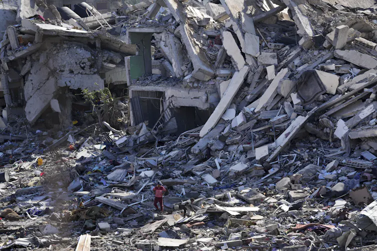 A huge pile of rubble and destroyed buildings dwarf people standing in front of them.