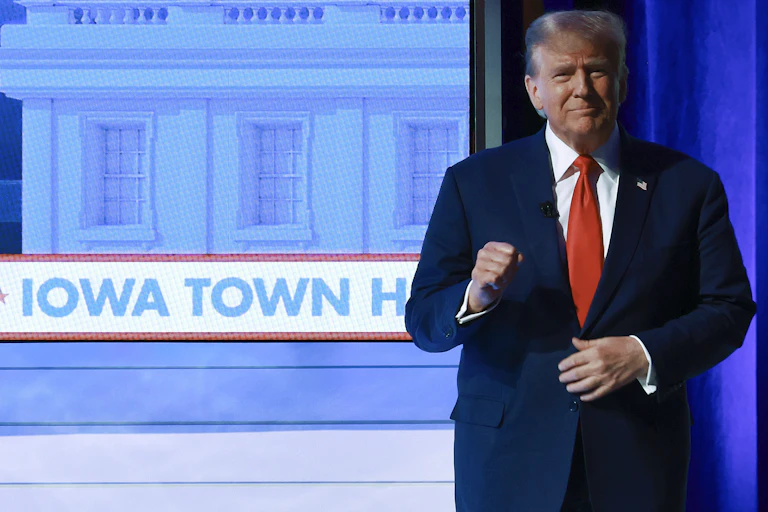 A gray-haired white man dressed in a dark suit with a white shirt and red tie stands in front of an image of the words Iowa Town Hall.
