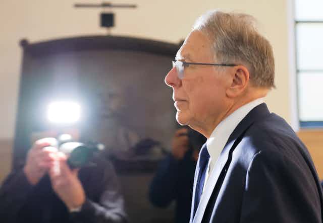 Man with gray hair and glasses in a suit looks straight ahead while someone takes his photo.