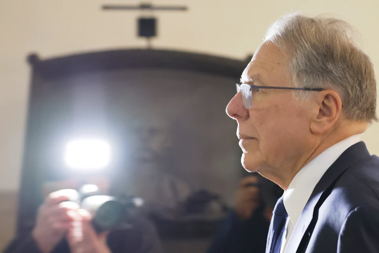 Man with gray hair and glasses in a suit looks straight ahead while someone takes his photo.