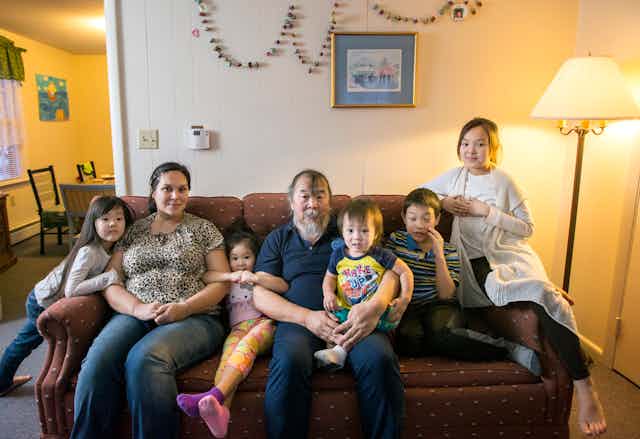 Two adults, including one man with a long white beard, sit on a couch with five small children. 