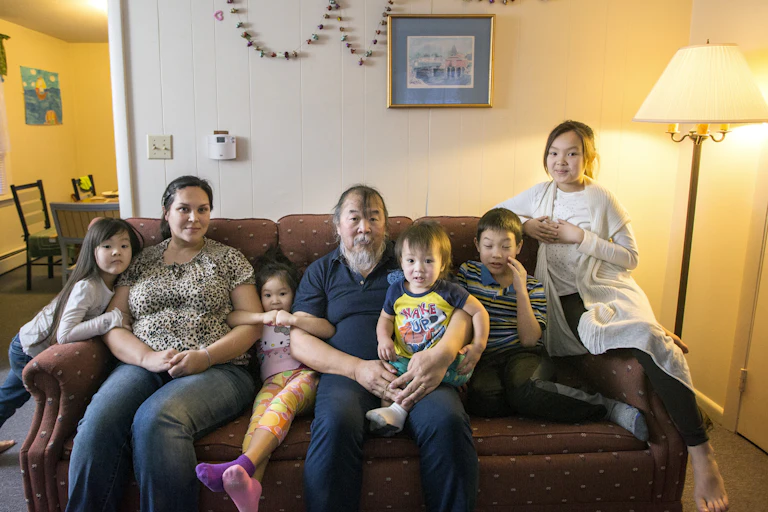 Two adults, including one man with a long white beard, sit on a couch with five small children.