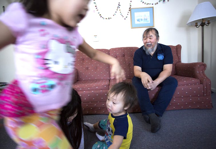 A man with a grey beard sits on a red couch and watches young children run around.