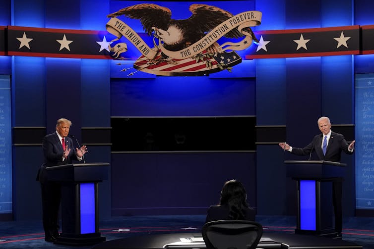 Two older men stand behind podiums several feet apart with blue drapery and lighting behind them.