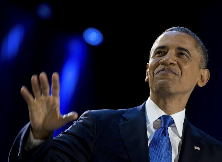 A Black man smiles and waves as he looks up from a stage.