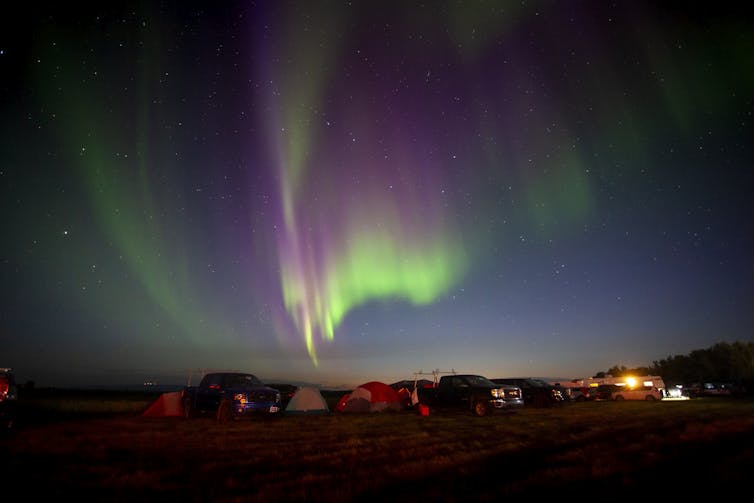 Tents and vehicles are seen in a field under a clear night sky filled with Aurora Borealis