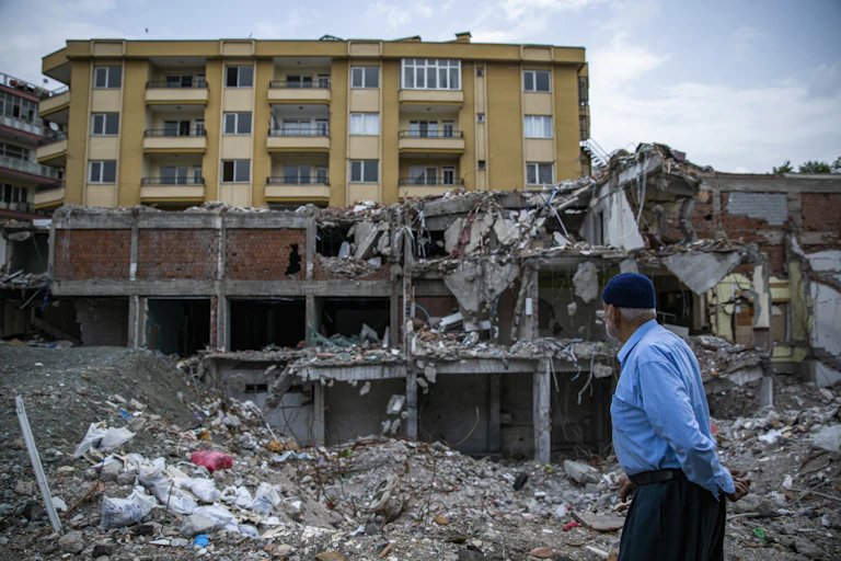 A man wearing a blue cap and shirt walks passed a destroyed building. Another still intact building can be seen behind it.