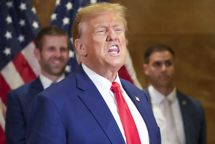 A blond-ish man in a blue suit and red tie grimaces while speaking, his tongue visible between his teeth.