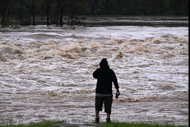 A man with his back to the camera stands in front of rushing floodwaters in Queensland