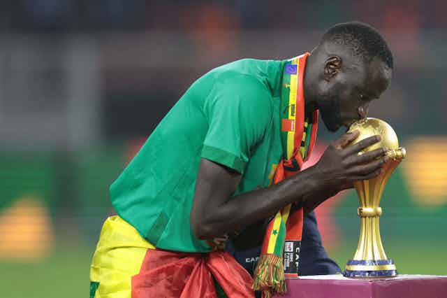A man in football gear with a scarf in the colours of the Senegalese flag bends and kisses a gold trophy of a football.