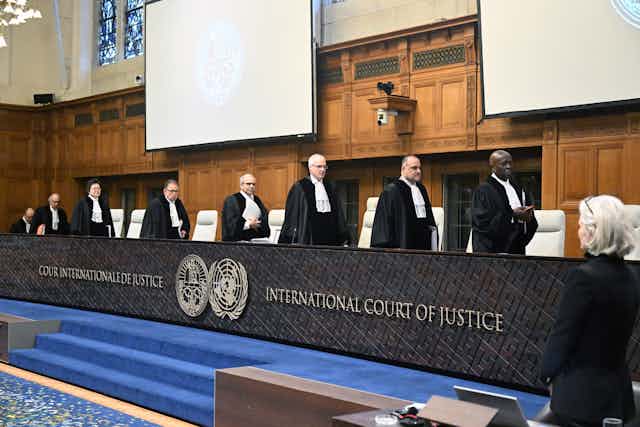 Eight people wearing dark robes and white scarflike ties walk toward a row of seats behind a black barrier that says 'International Court of Justice.'
