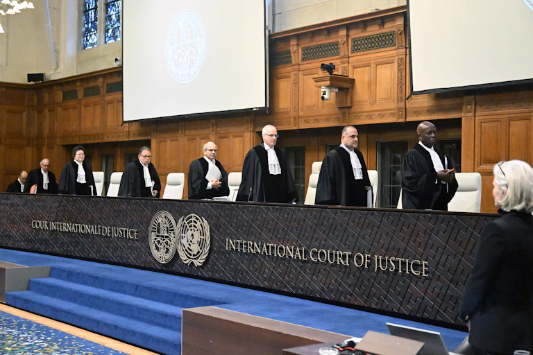 Eight people wearing dark robes and white scarflike ties walk toward a row of seats behind a black barrier that says 'International Court of Justice.'