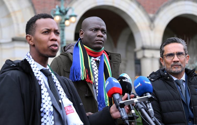 Three men wear scarves, including one with red, blue and green, stand in front of a set of microphones and look past the camera.