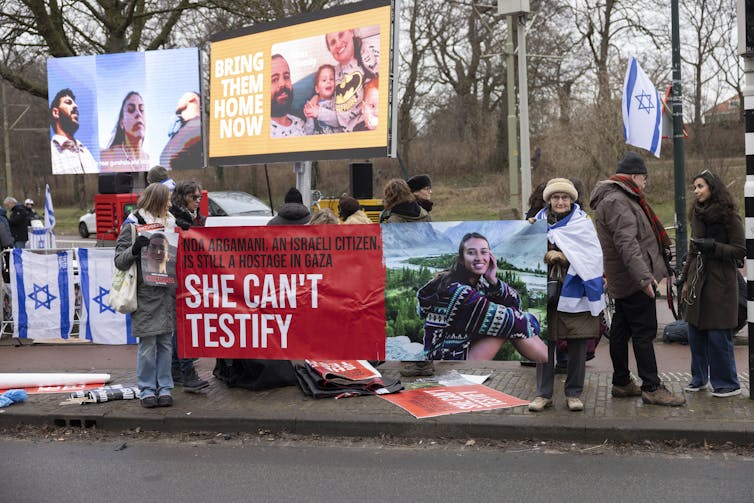 People stand on the street and hold up large banners of kidnapped Israeli family members, with the words 'Bring them home now,' and 'She can't testify.' They also have Israeli flags.