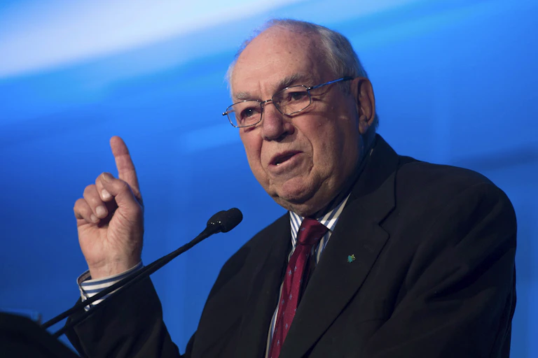 An older man in rectangular glasses raises his finger while speaking into a microphone